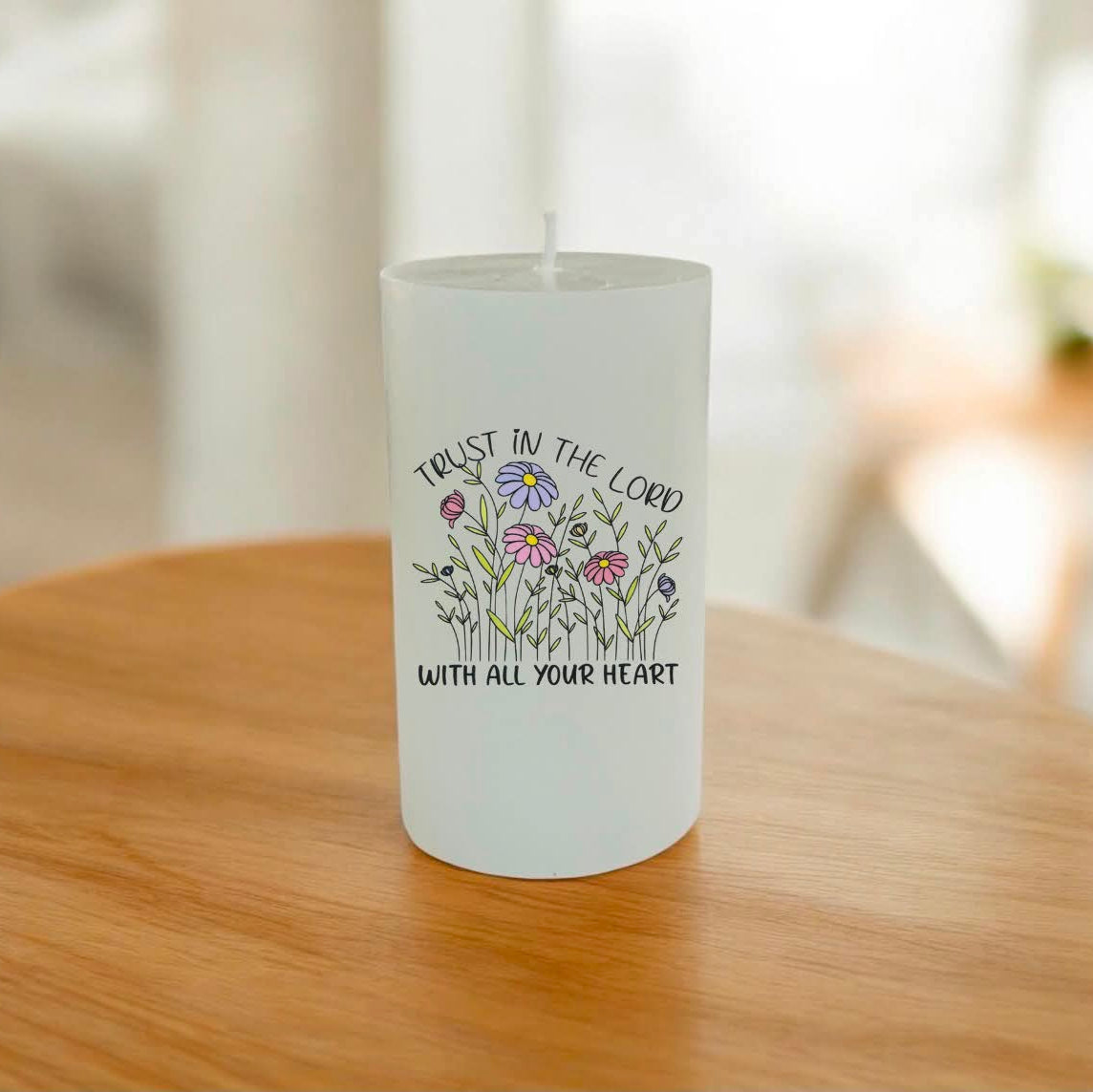 A white pillar candle with a floral illustration and the printed words 'TRUST IN THE LORD WITH ALL YOUR HEART' sits on a wooden table in a softly blurred indoor setting.