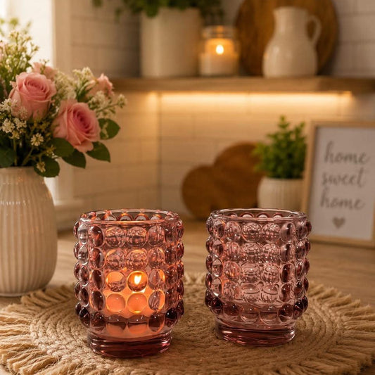 Two textured, bubble-patterned glass candle holders in a dusty rose pink color, on a wooden table in a cozy, warmly lit kitchen setting.