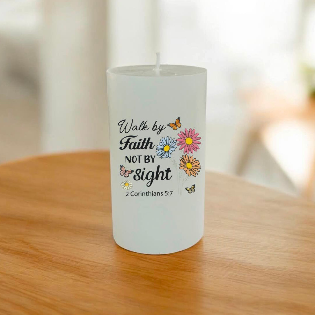 A white pillar candle with a printed quote reading 'Walk by Faith NOT BY sight' and small colourful flowers and butterflies, sitting on a wooden table in a softly blurred indoor setting.