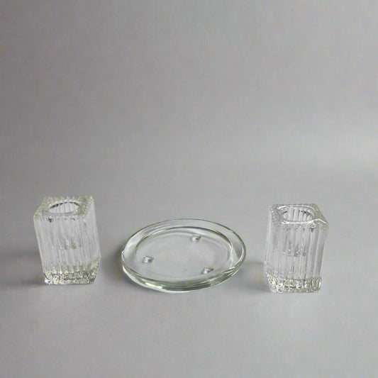 Two clear glass candle holders and a round glass lid on a white surface with a blurred background of shelves.