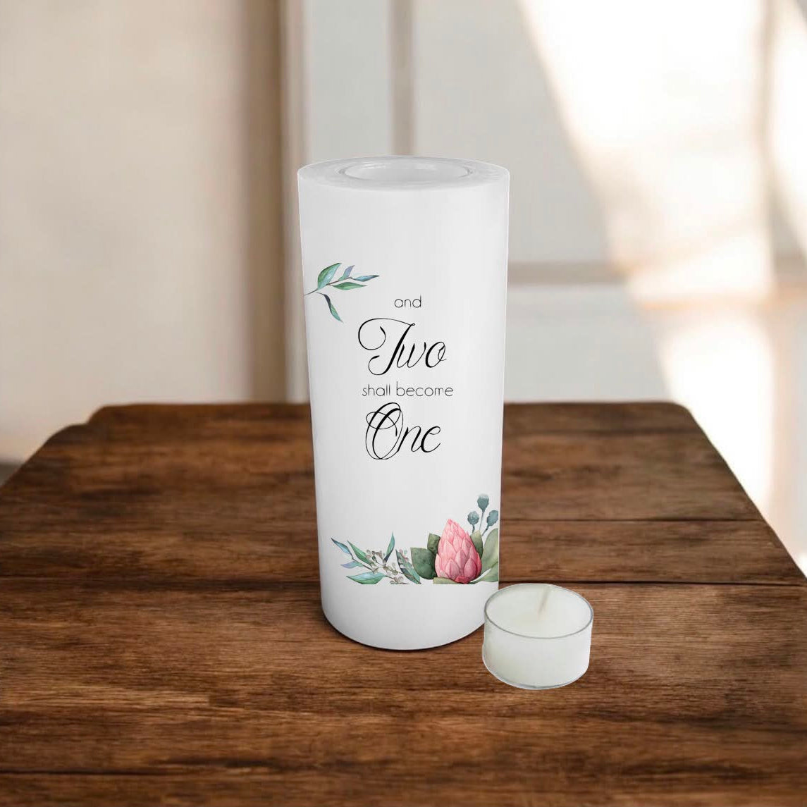 A white candle with printed script text reading 'and two shall become one' and delicate floral illustrations, displayed on a rustic wooden surface.