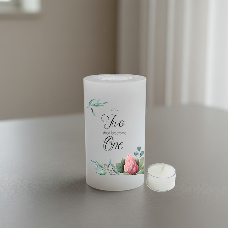 A white pillar candle with printed script text reading 'and Two shall become One' and small floral illustrations in green and pink, shown on a tabletop in a softly lit indoor setting.