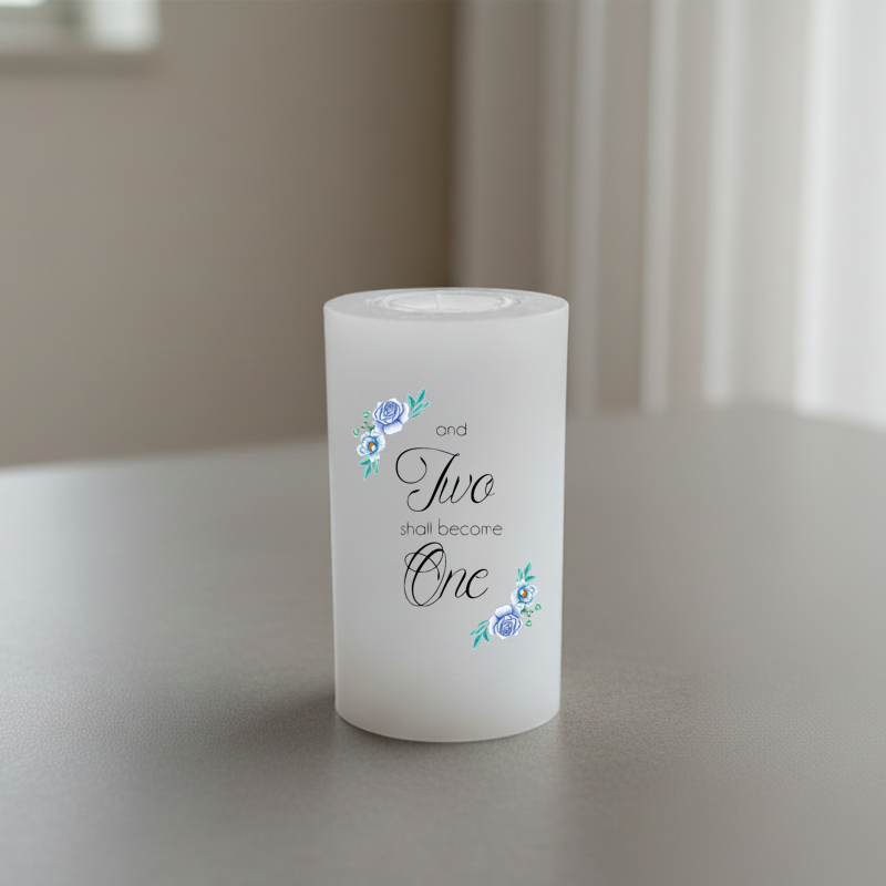 A white candle with printed text reading 'and Two shall become One' in black script and serif fonts, accented with small blue and green floral illustrations.