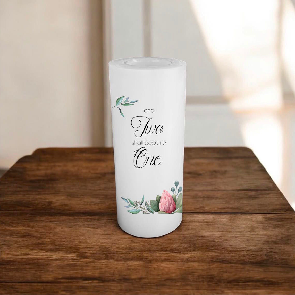 A white candle with printed script text reading 'and two shall become one' and delicate floral illustrations, displayed on a rustic wooden surface.