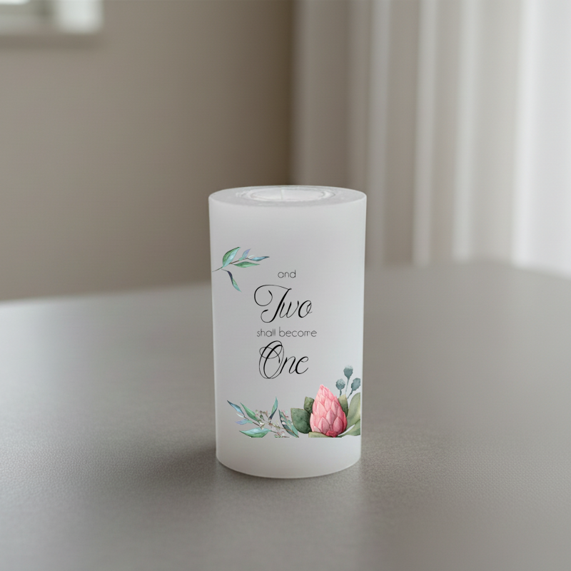A white pillar candle with printed script text reading 'and Two shall become One' and small floral illustrations in green and pink, shown on a tabletop in a softly lit indoor setting.
