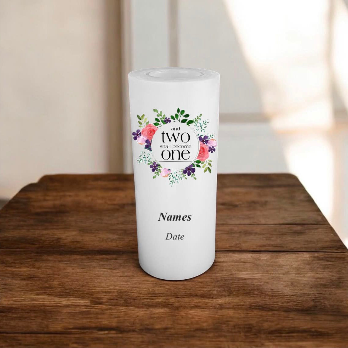 A white candle with a printed text 'and TWO shall become ONE' inside a floral wreath, shown on a wooden table.