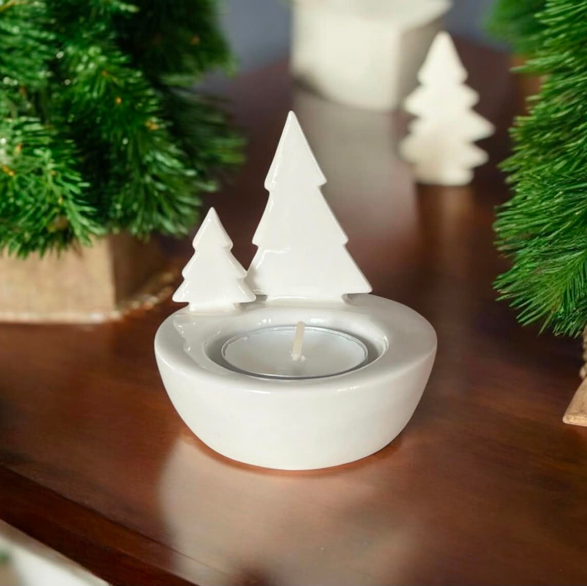 A white ceramic tea light candle holder shaped like two small Christmas trees, displayed on a dark wooden surface with green pine branches in the background.