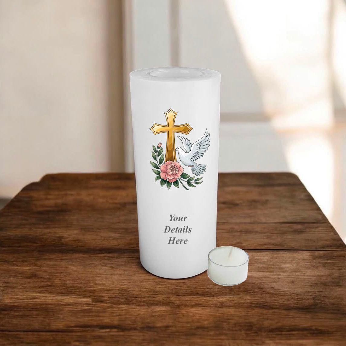White candle with a printed gold cross, white dove, pink rose, and green leaves, shown on a wooden table next to a small white tea light candle.