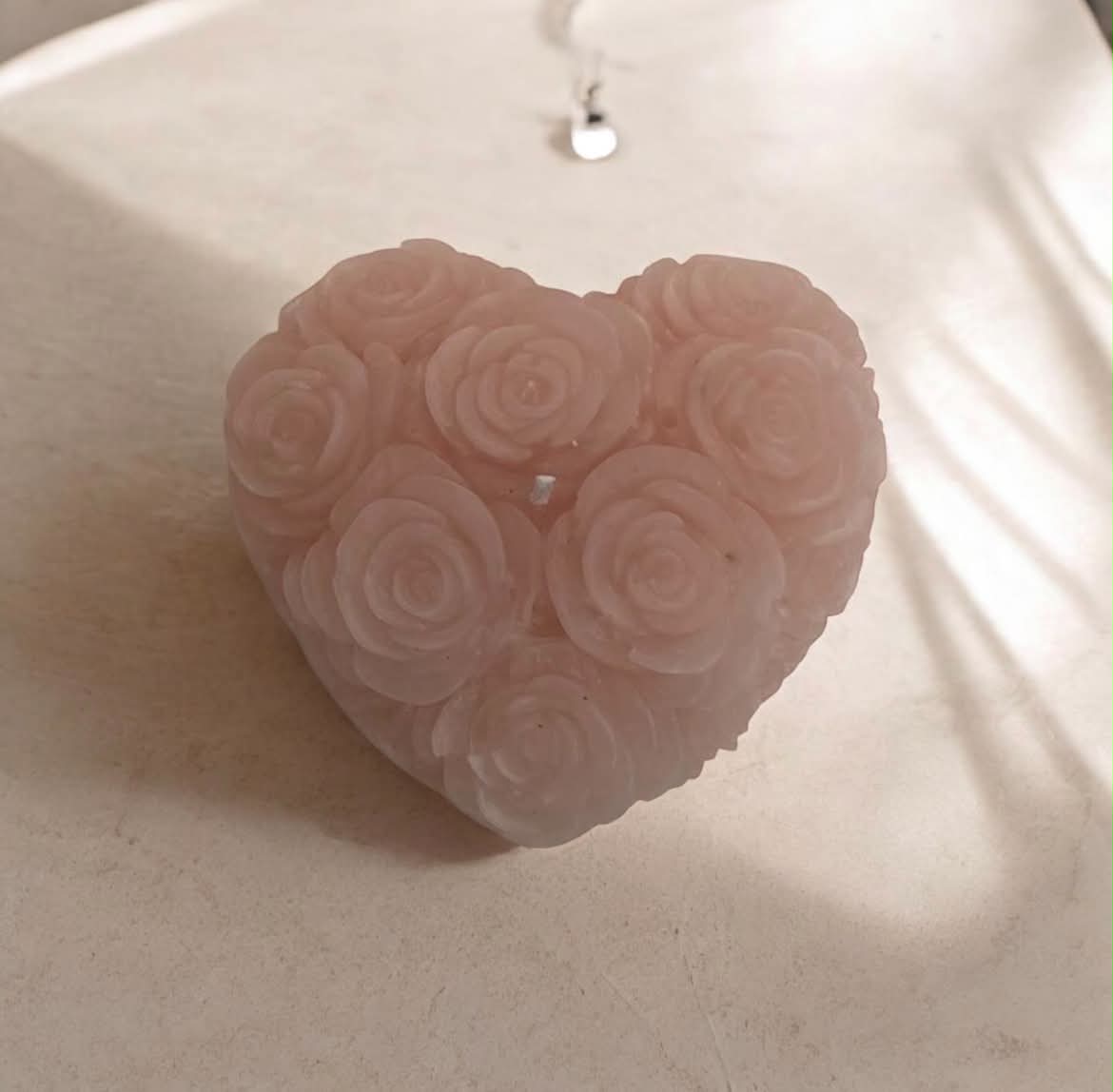 A heart-shaped rose wax melt or candle with a carved rose pattern, shown on a neutral beige surface with a silver pendant necklace in the background.
