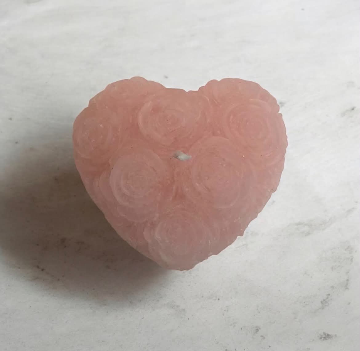 A heart-shaped pink soap or candle featuring a carved rose pattern, resting on a light gray marble surface.