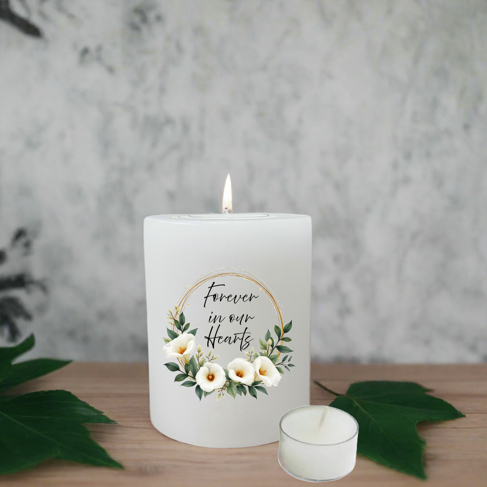 A white candle with a single lit flame stands on a wooden surface, decorated with a floral wreath and the words 'Forever in our Hearts.' A smaller unlit tealight candle sits in front.