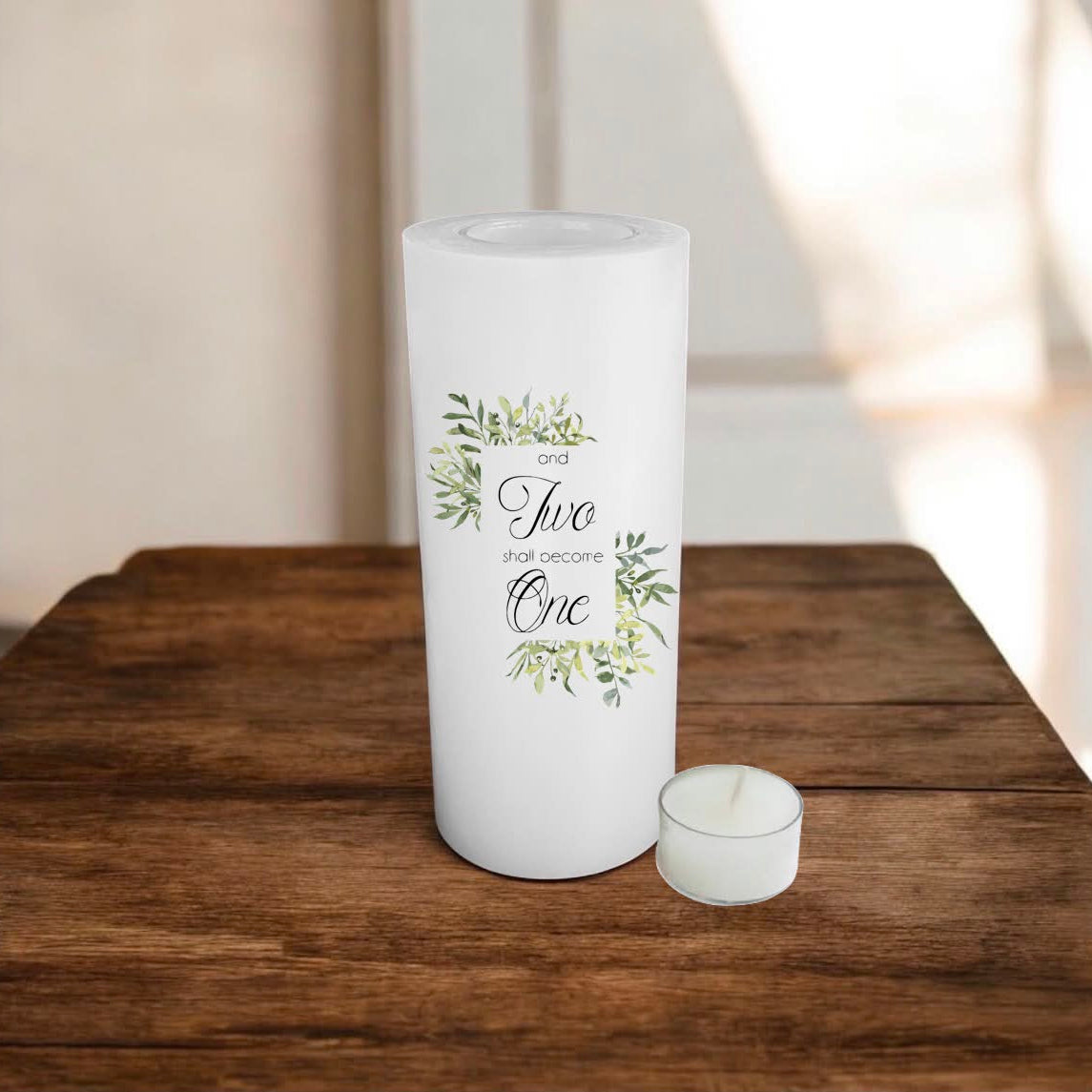 A white candle holder with a green flowery design and the text. Sits on a rustic wooden table with a softly blurred indoor background.