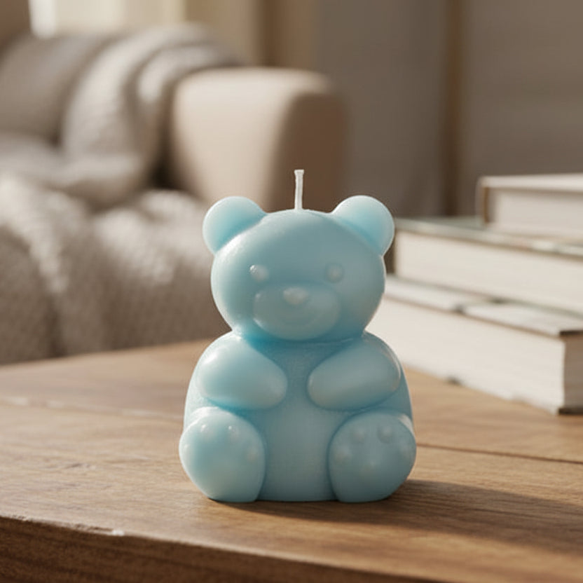 A small light-blue bear-shaped candle with a wick on top, standing on a coloured surface with a blurred background.