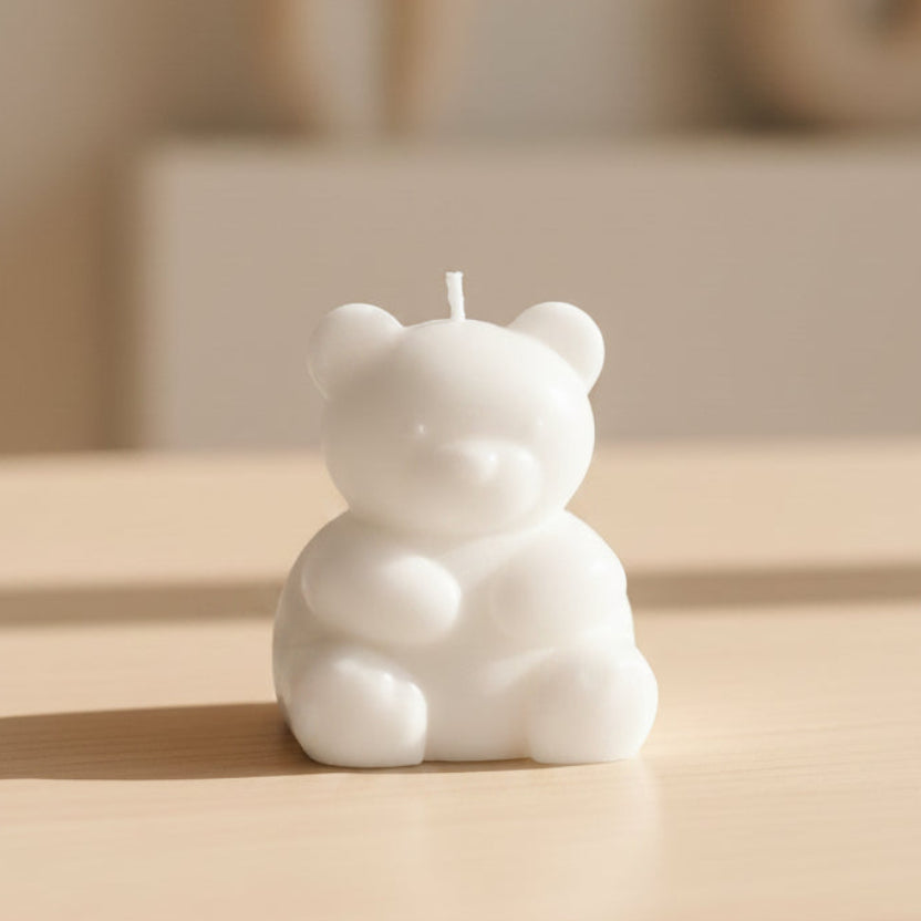 A small white bear-shaped candle with a wick on top, standing on a coloured surface with a blurred background.