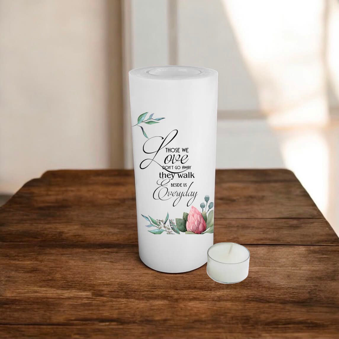 A white candle with a printed quote and floral illustration, standing on a rustic wooden surface against a softly blurred indoor background.