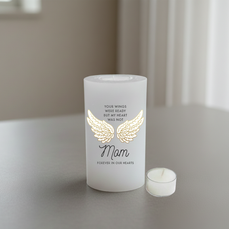 A white memorial candle with a gold angel wings graphic and the word 'Mom' in black script.