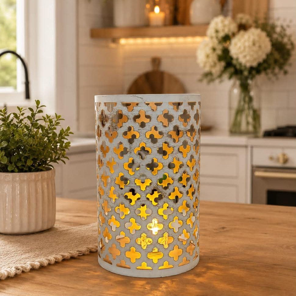 A cylindrical metal lantern with a white cutout geometric pattern and warm yellow-gold interior lighting, placed on a wooden table in a bright kitchen setting.