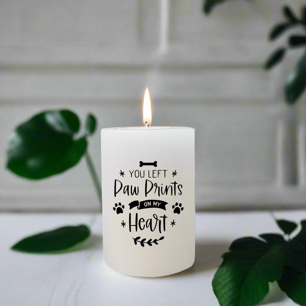 White candle with 'You Left Paw Prints on My Heart' text on a white surface with green leaves.