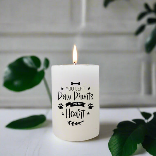 White candle with 'You Left Paw Prints on My Heart' text on a white surface with green leaves.