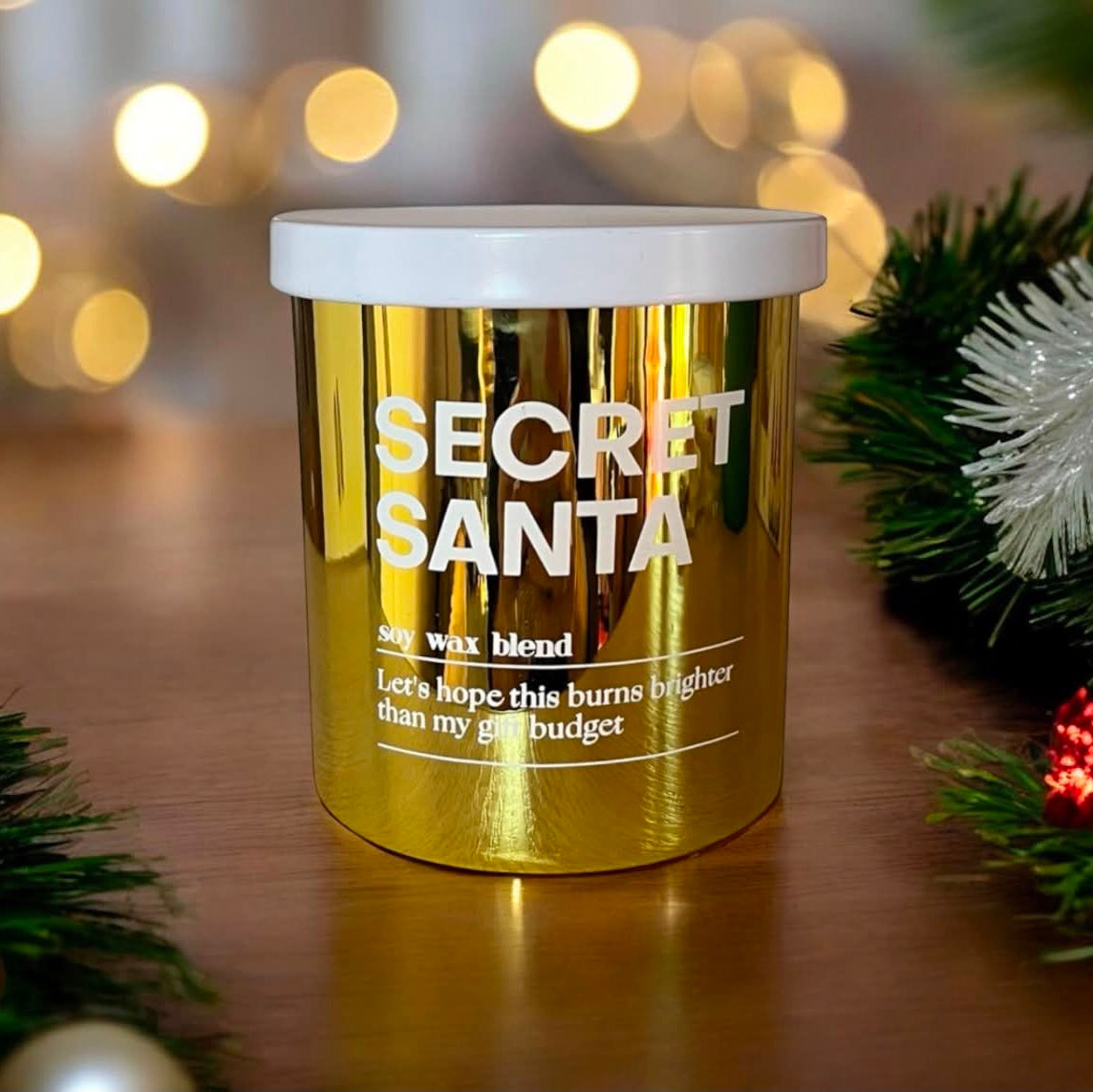 A shiny gold jar candle with a white lid labeled 'SECRET SANTA' sits on a wooden surface with Christmas greenery and warm blurred lights in the background.