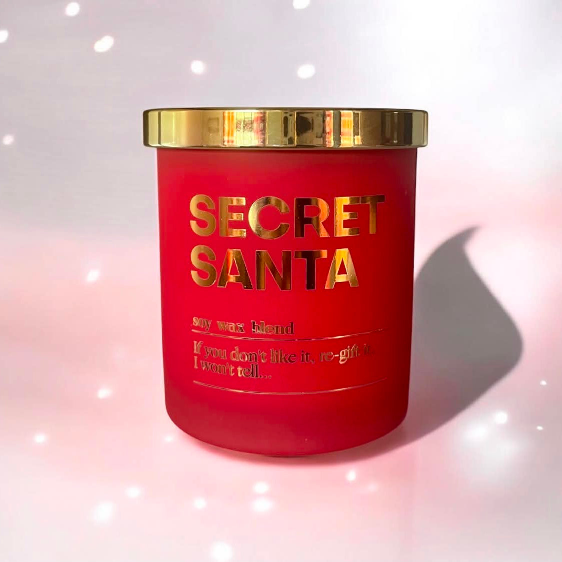 A red cylindrical candle jar with a shiny gold lid and gold lettering that reads 'SECRET SANTA' on the front.