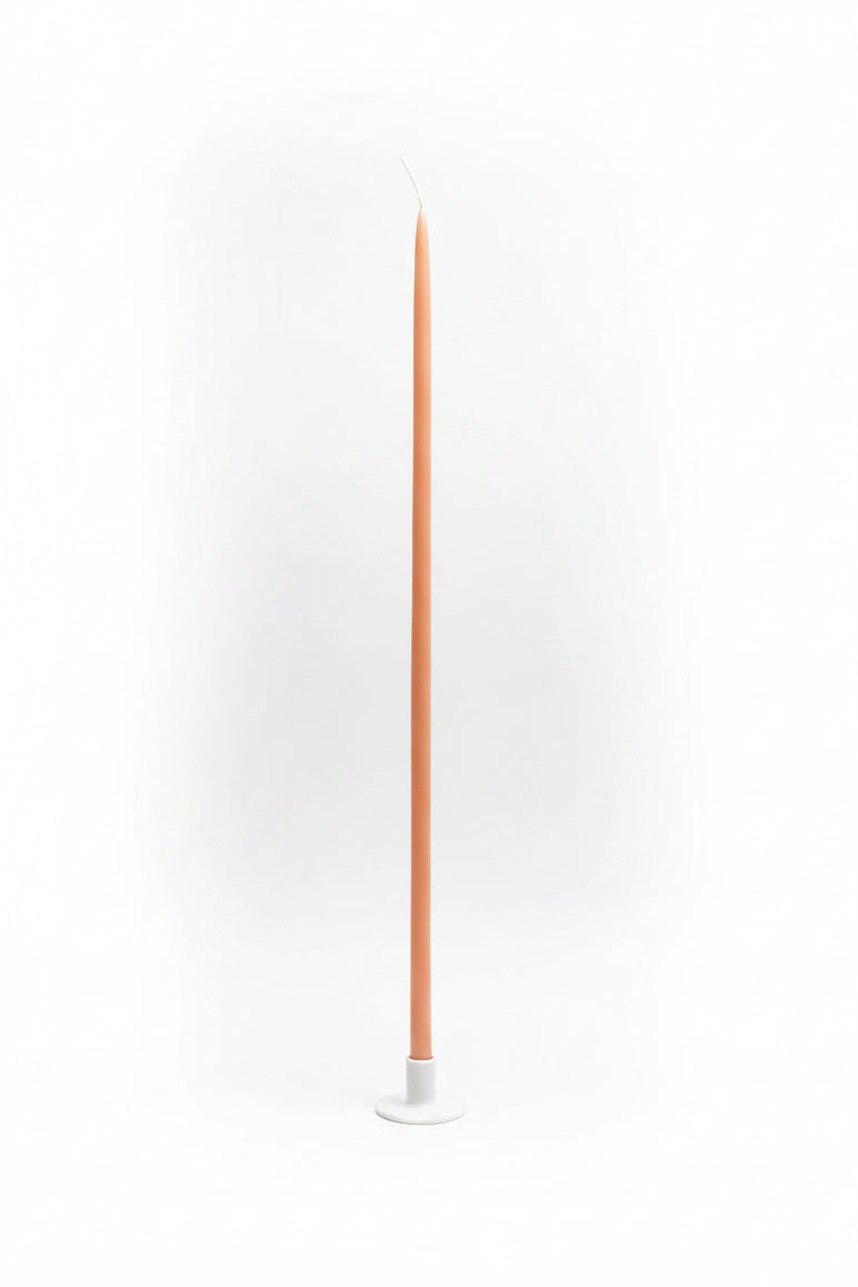 A tall, slender peach taper candle with a white base and a white loop at the top, standing upright on a white surface.