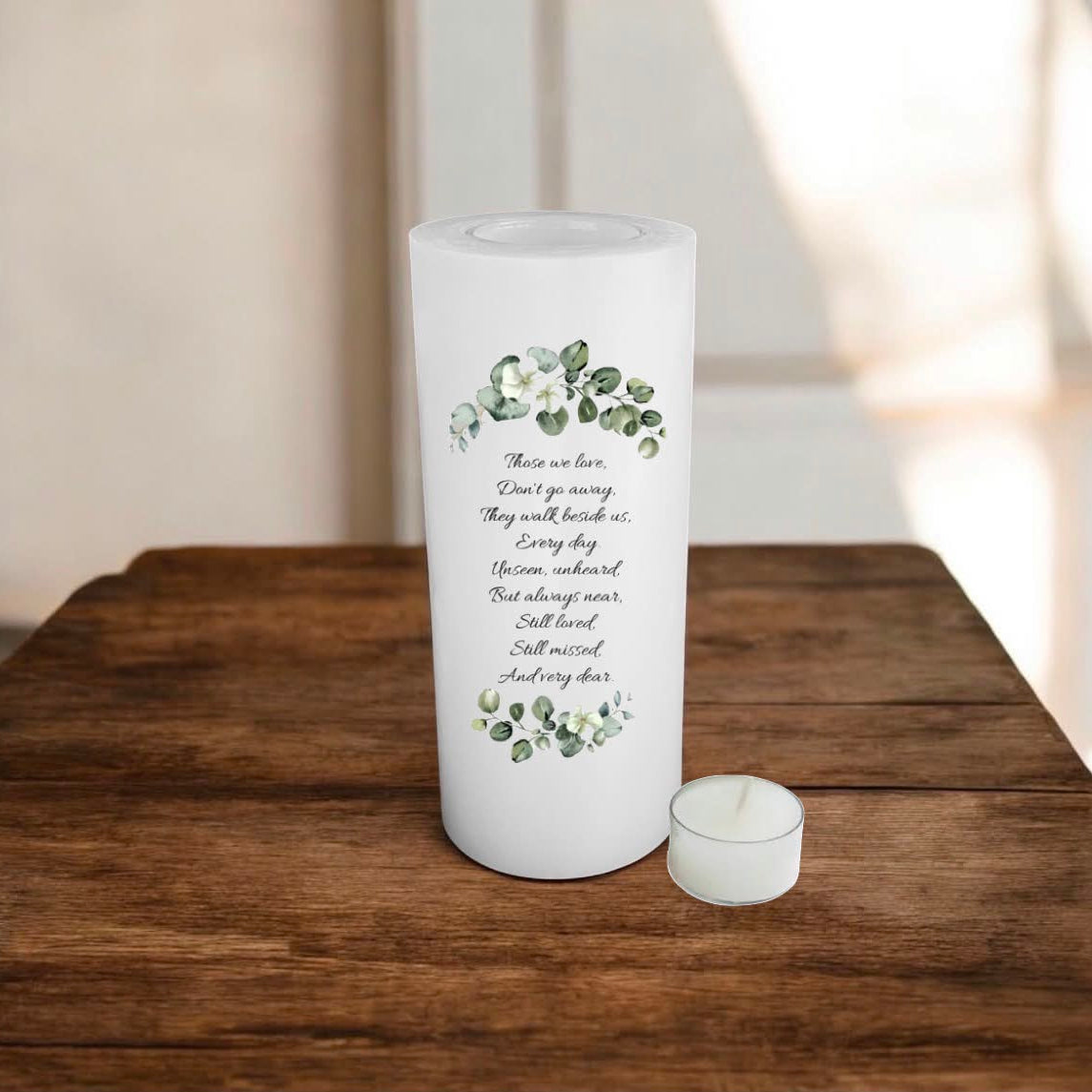 A white candle with printed black script text and green floral illustrations, standing on a wooden surface against a softly blurred indoor background.