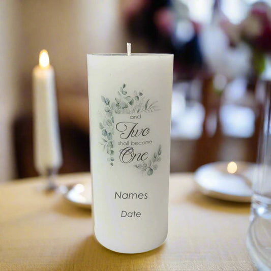 A white wedding candle with a lit wick stands on a table. The front of the candle is printed with a delicate green floral illustration and text