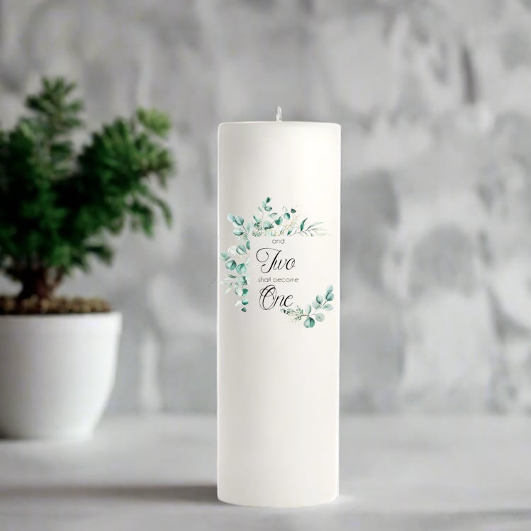 White candle with 'and two shall become one' text and floral design, placed on a surface with a blurred background.