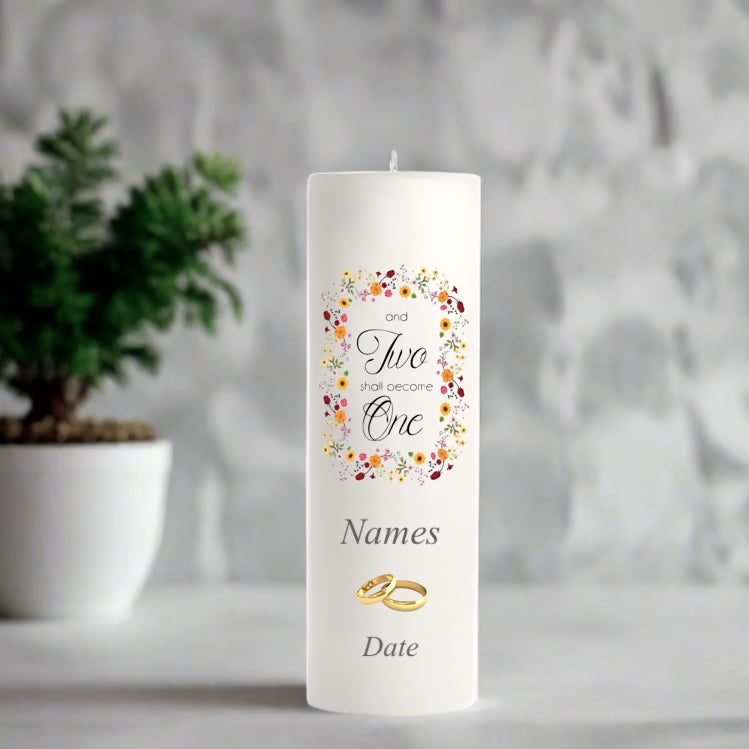 White wedding candle with 'and two shall become one' text and floral design, placed on a surface with a blurred background.