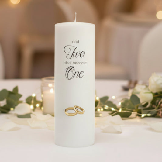 White wedding candle with 'Two shall become One' text and wedding rings, surrounded by flowers and lights.