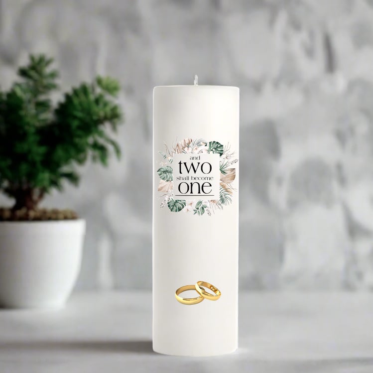 White wedding candle with floral design and gold rings, placed on a surface with a blurred plant and gray background.