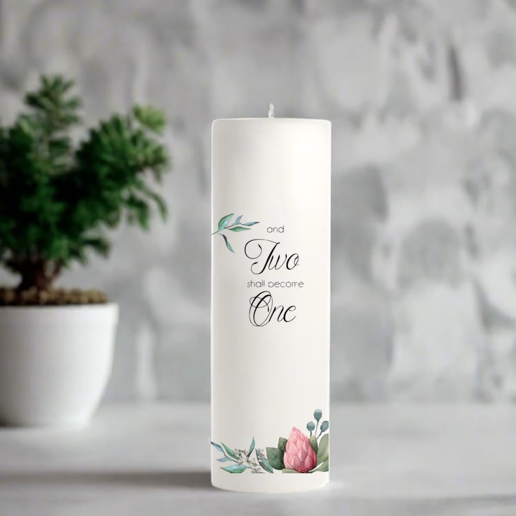 White wedding candle with floral design and text on a gray surface with a plant in the background