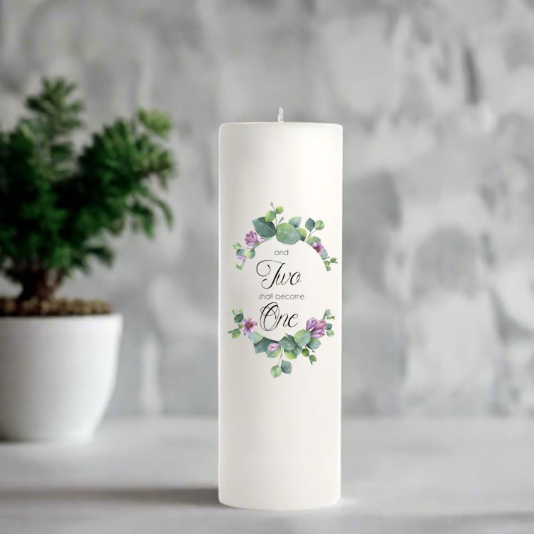 White wedding candle with floral design and text on a gray surface with a plant in the background