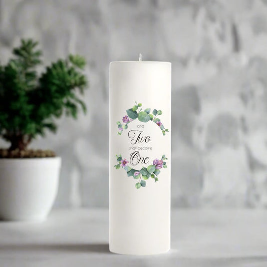White wedding candle with floral design and text on a gray surface with a plant in the background