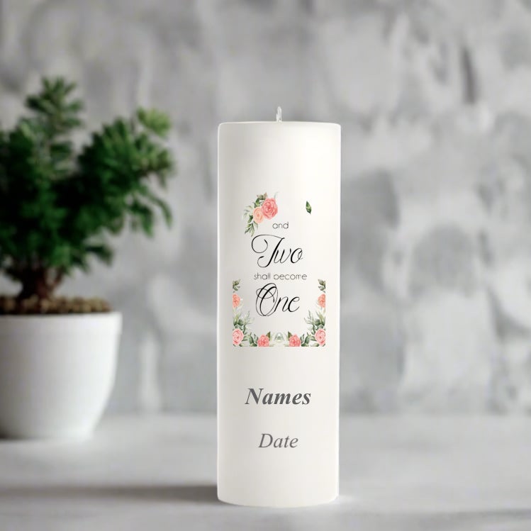 White wedding candle with floral design and text on a gray surface with a plant in the background