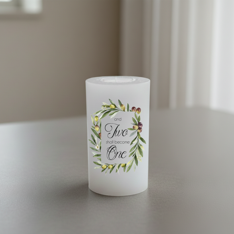 A white candle with a botanical wreath design and the printed phrase “and Two shall become One,” shown on a tabletop in a softly blurred indoor setting.