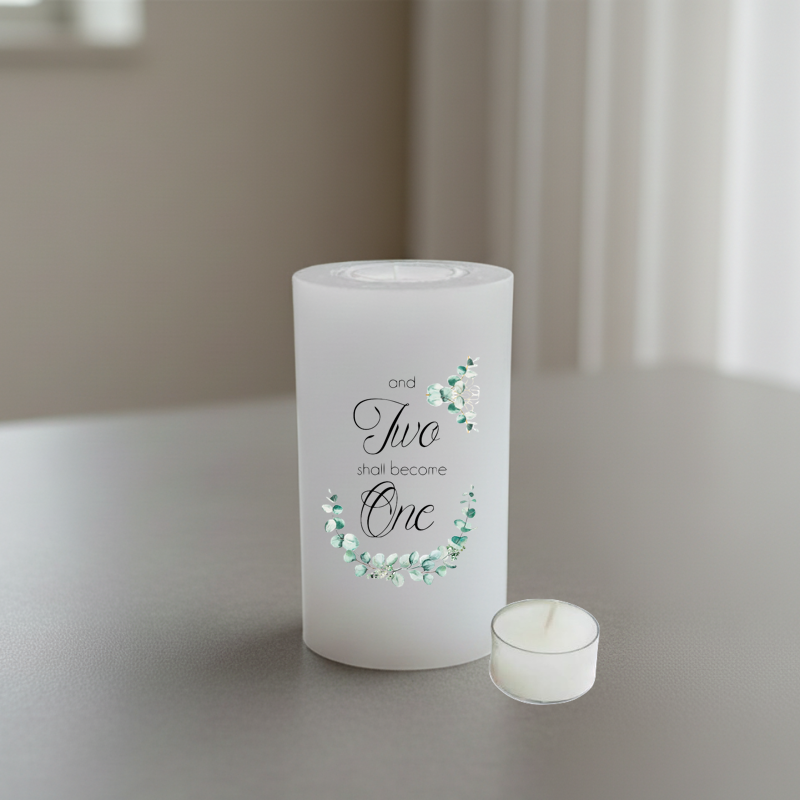 A white candle with printed text reading 'and Two shall become One' in black script and serif fonts, accented with green floral illustrations.
