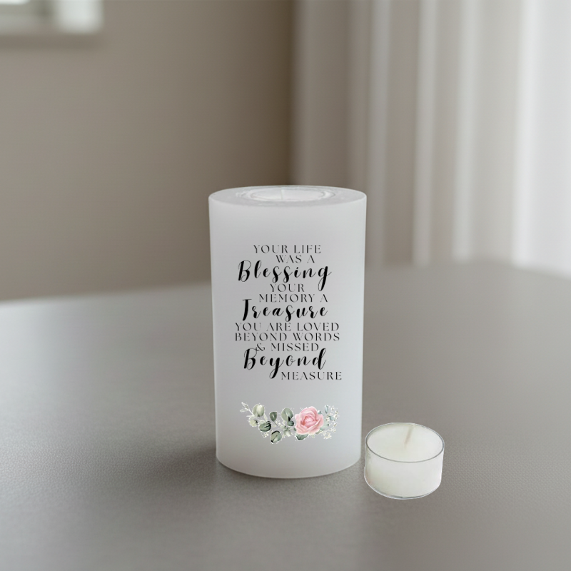 A white candle with black script text reading 'YOUR LIFE WAS A blessing YOUR MEMORY A Treasure YOU ARE LOVED BEYOND WORDS & MISSED Beyond MEASURE' with a small pink and green floral illustration.