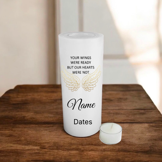 A white memorial candle with printed text and angel wings design sits on a wooden table